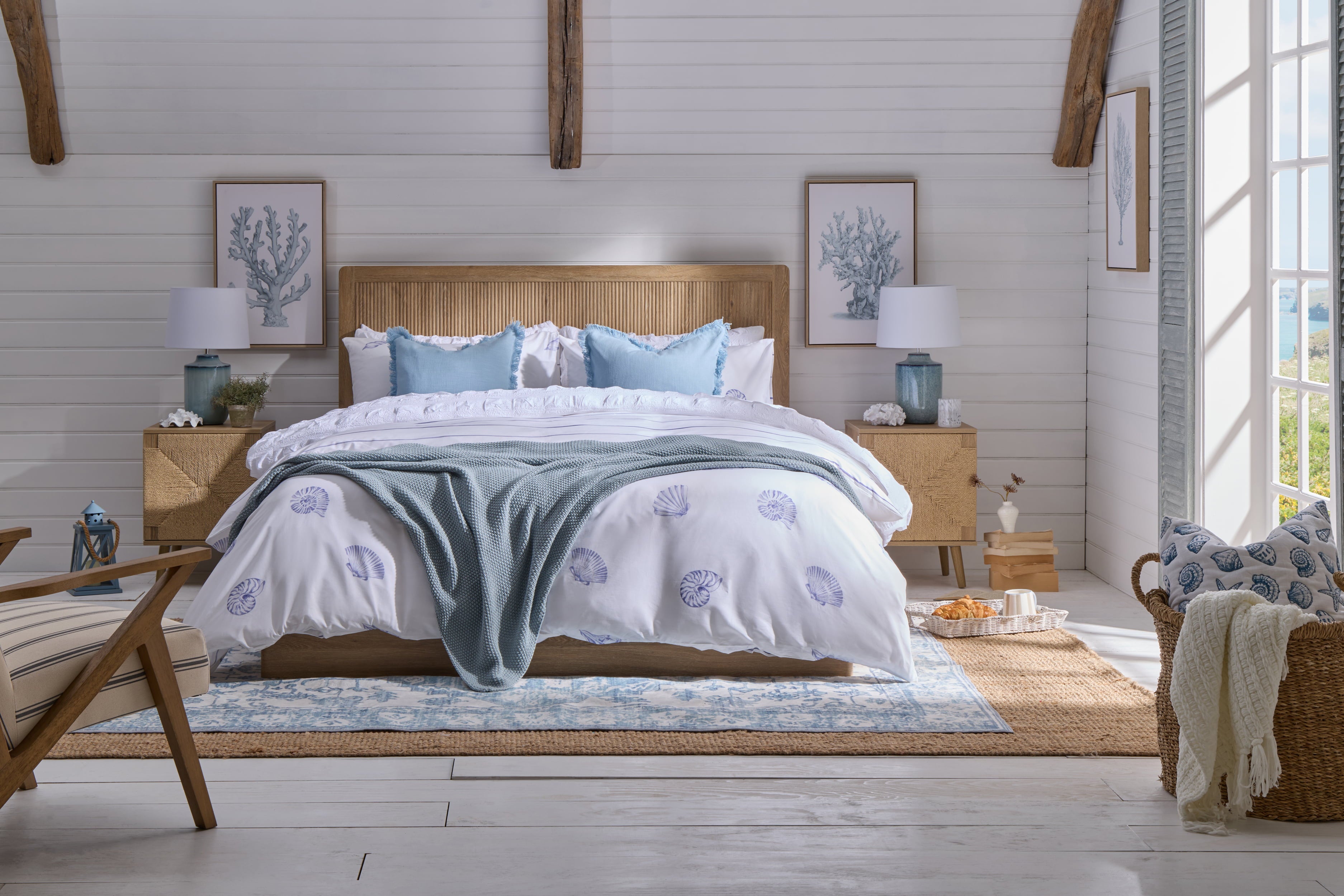 Coastal bedroom with shell print bedding, blue throw, light wood furniture and woven bedside tables