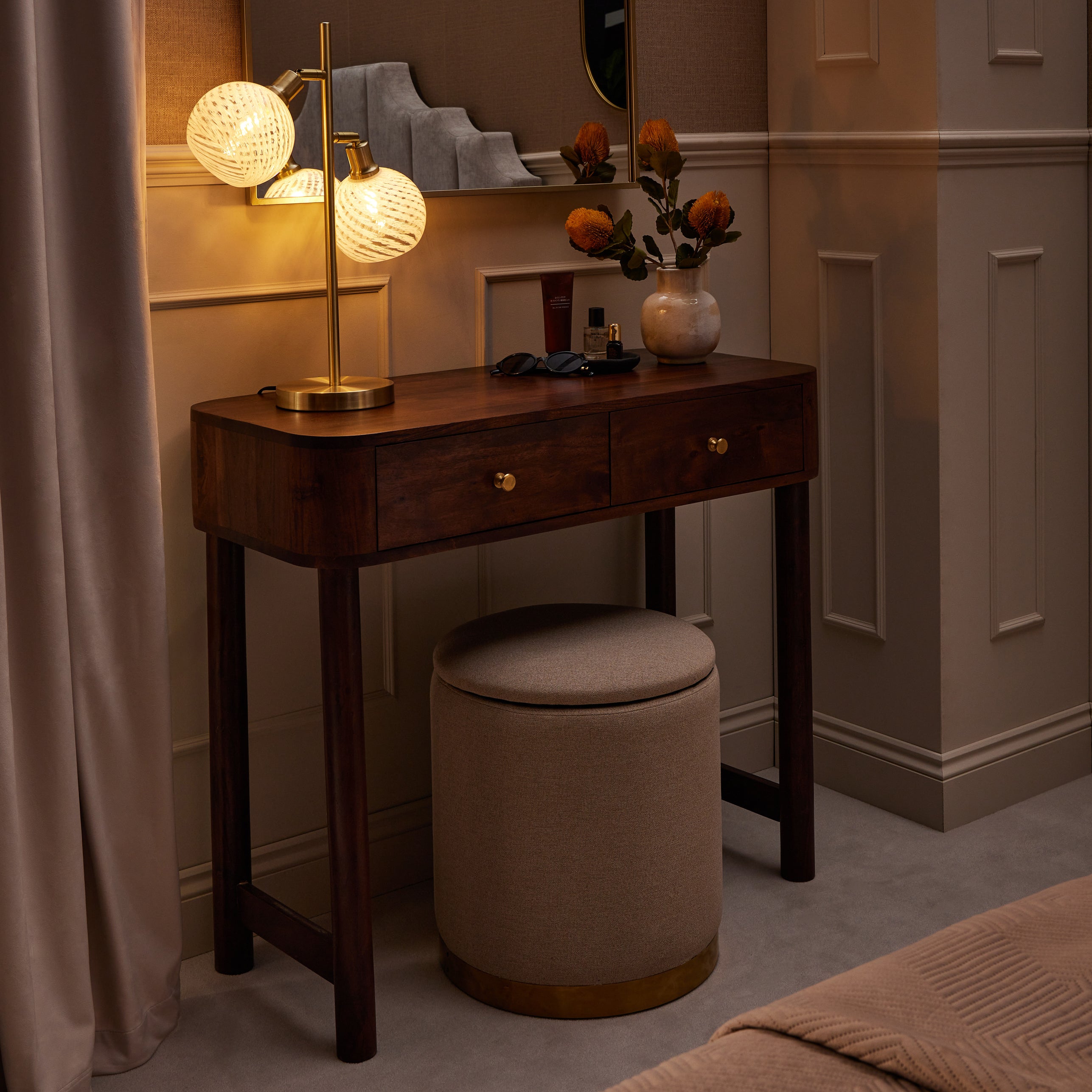 A wooden dressing table styled with a brass double globe lamp, neutral stool, and decorative accessories against panelled taupe walls.
