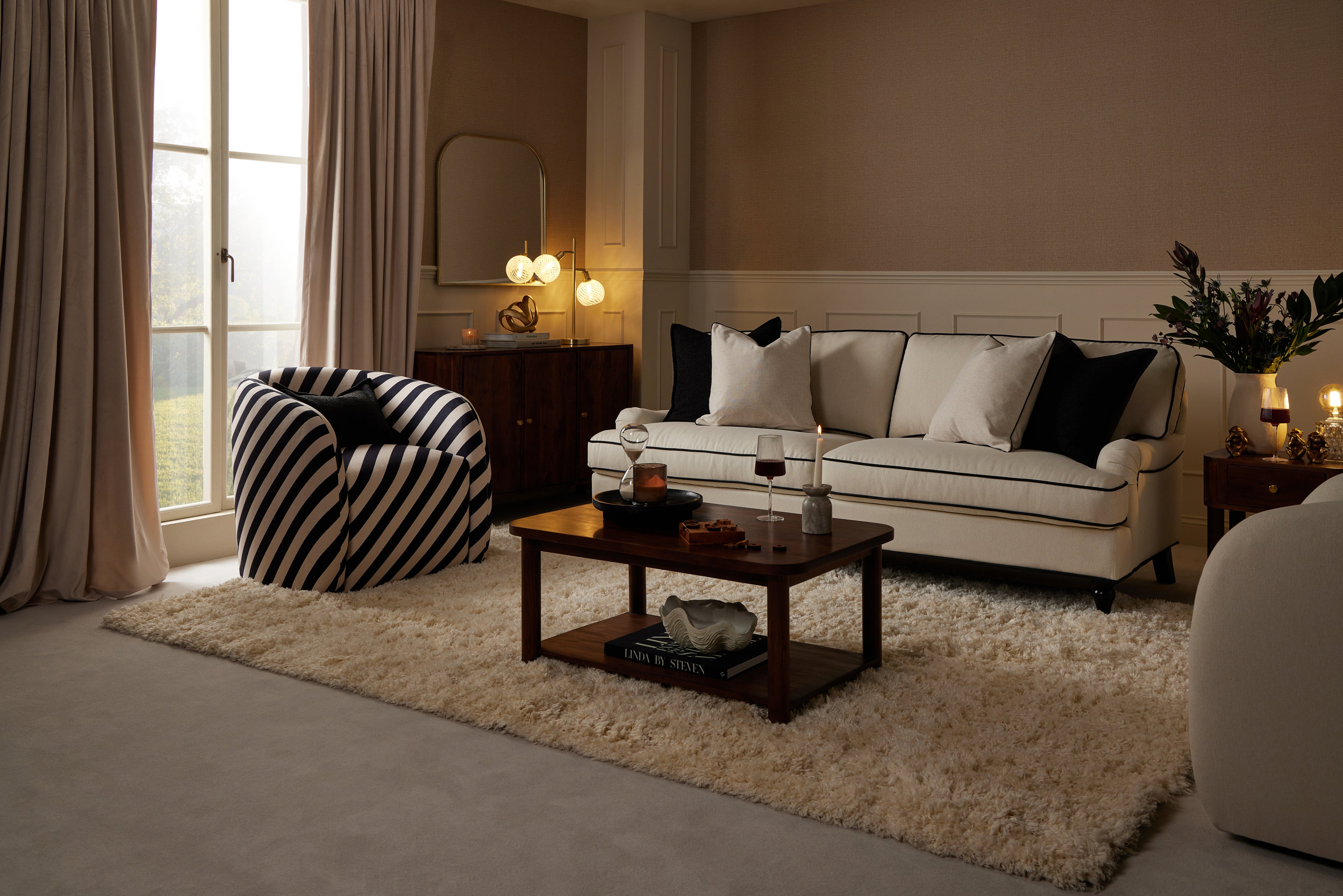 A cosy living room with a cream sofa, striped accent chair, shaggy rug, and dark wood coffee table, styled with soft warm lighting.