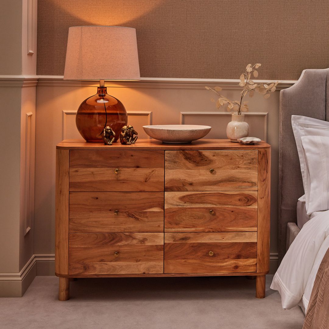A natural wood chest of drawers styled with a warm amber lamp, decorative bowl, and vase of stems beside a neutral upholstered bed.